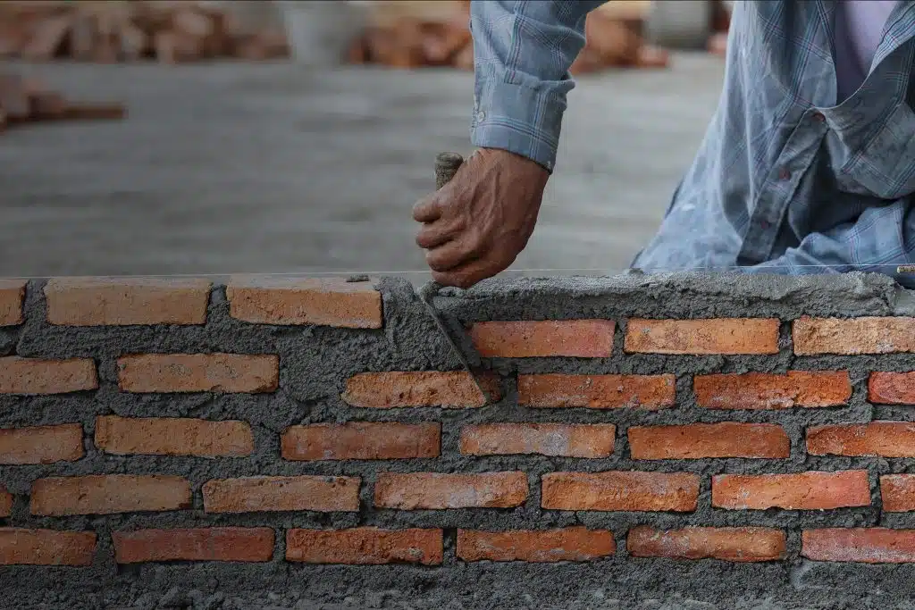This image shows the hands of a mason or bricklayer working on constructing a brick wall The image focuses on the workers hands which are calloused and covered in mortar as they carefully place a brick into the wall The wall is made of red and brown bricks that are laid in a traditional pattern with mortar visible between the bricks The background is slightly blurred but appears to be a construction site or outdoor area
