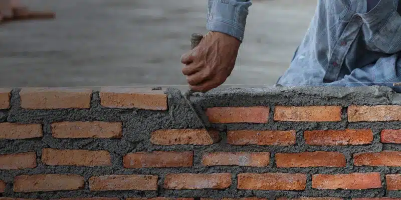 This image shows the hands of a mason or bricklayer working on constructing a brick wall. The image focuses on the worker's hands, which are calloused and covered in mortar, as they carefully place a brick into the wall. The wall is made of red and brown bricks that are laid in a traditional pattern, with mortar visible between the bricks. The background is slightly blurred, but appears to be a construction site or outdoor area.
