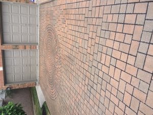 This image shows a paved driveway or walkway in front of a brick building The paving consists of interlocking brick pavers arranged in a circular pattern at the center surrounded by a grid-like pattern of rectangular bricks The bricks have a warm tan-colored tone and are laid in a visually appealing and symmetrical design The image highlights the masonry work and attention to detail in the paving which appears to be a well-crafted and aesthetically pleasing feature of the property