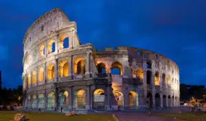 The image shows the iconic Colosseum in Rome Italy illuminated at night The massive ancient Roman amphitheater is made of stone and features numerous arched entrances and windows The exterior walls are lit up creating a warm golden glow that contrasts with the deep blue of the night sky The Colosseums distinctive architecture and grandeur are prominently displayed showcasing its historical significance and architectural beauty