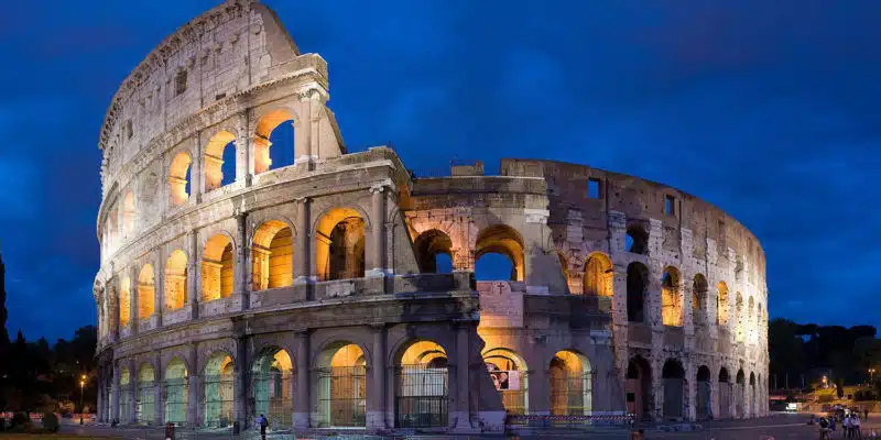 The image shows the iconic Colosseum in Rome, Italy, illuminated at night. The massive ancient Roman amphitheater is made of stone and features numerous arched entrances and windows. The exterior walls are lit up, creating a warm, golden glow that contrasts with the deep blue of the night sky. The Colosseum's distinctive architecture and grandeur are prominently displayed, showcasing its historical significance and architectural beauty.