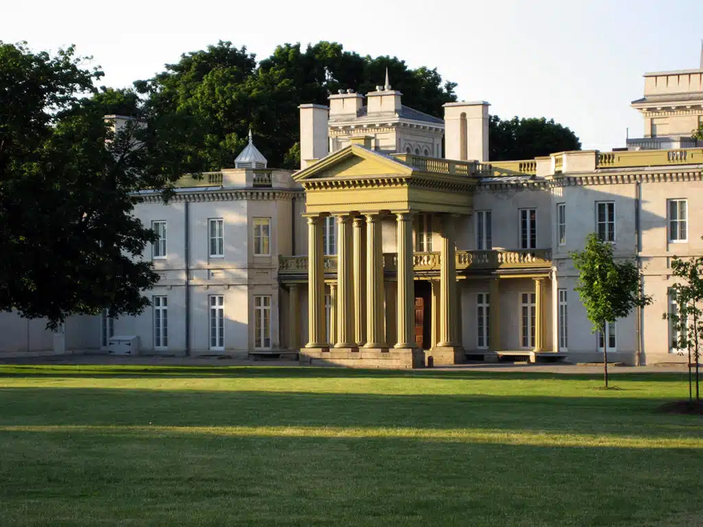 This image depicts a grand historic building with a classical architectural style The building has a prominent central portico with columns and a pediment surrounded by wings on either side The exterior is a light neutral color with ornate details and decorative elements The building is set against a backdrop of lush green trees and is situated on a well-manicured lawn The overall scene conveys a sense of grandeur and stately elegance