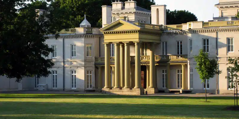 This image depicts a grand, historic building with a classical architectural style. The building has a prominent central portico with columns and a pediment, surrounded by wings on either side. The exterior is a light, neutral color with ornate details and decorative elements. The building is set against a backdrop of lush, green trees, and is situated on a well-manicured lawn. The overall scene conveys a sense of grandeur and stately elegance.