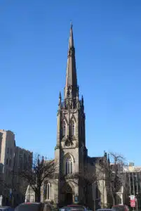 This image shows a tall Gothic-style church steeple against a clear blue sky The steeple has a distinctive spire shape with intricate architectural details including pointed arches ornate carvings and a weather vane on top The church building itself is not fully visible in the frame but the steeple dominates the scene rising high above the surrounding urban landscape