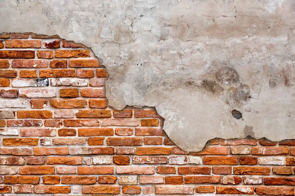 This image shows a textured wall surface composed of a mix of exposed brick and plaster The brick portion features rows of reddish-orange bricks some of which are partially crumbling or damaged creating an aged and weathered appearance The plaster area has a rough uneven texture with visible cracks and discoloration giving the overall wall a distressed vintage aesthetic The contrast between the brick and plaster creates an interesting visual texture and a sense of the walls history