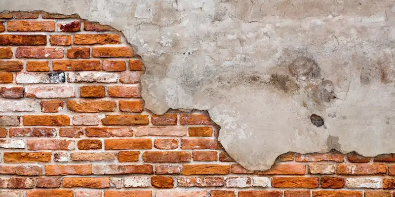 This image shows a textured wall surface composed of a mix of exposed brick and plaster. The brick portion features rows of reddish-orange bricks, some of which are partially crumbling or damaged, creating an aged and weathered appearance. The plaster area has a rough, uneven texture with visible cracks and discoloration, giving the overall wall a distressed, vintage aesthetic. The contrast between the brick and plaster creates an interesting visual texture and a sense of the wall's history.