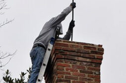 This image shows a worker, wearing a gray jacket and jeans, repairing or maintaining a brick chimney or structure. The worker is using a tool or equipment to work on the brickwork, which appears to be an older, weathered brick structure. The image conveys the skilled labor and attention to detail required for masonry work.