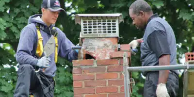 This image shows two masonry workers repairing a brick chimney or structure outdoors The workers are wearing protective gear like gloves and hard hats and are using tools and materials to work on the brickwork The scene is set in a wooded natural environment with lush green foliage in the background The image conveys the skilled hands-on work involved in masonry repairs and construction