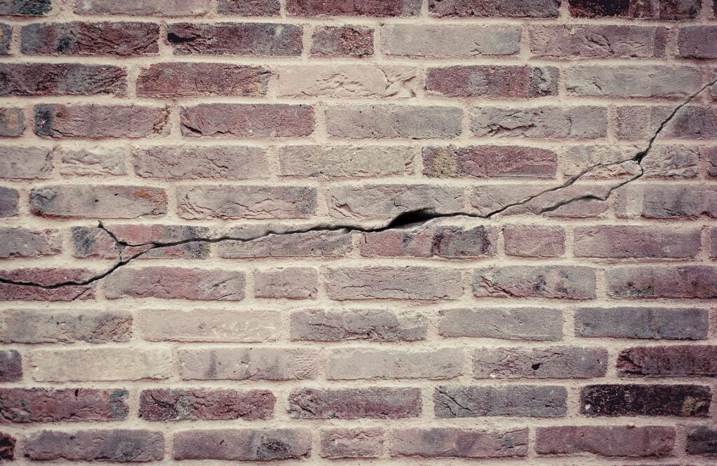 This image shows a textured brick wall The bricks are a mix of reddish-brown and gray tones with varying shades and textures The wall has a weathered aged appearance with visible cracks and crevices throughout the brickwork The overall impression is of a sturdy rustic masonry structure
