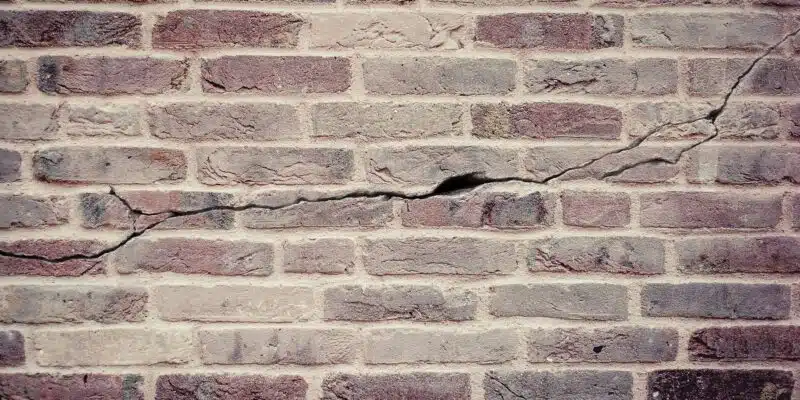 This image shows a textured brick wall. The bricks are a mix of reddish-brown and gray tones, with varying shades and textures. The wall has a weathered, aged appearance, with visible cracks and crevices throughout the brickwork. The overall impression is of a sturdy, rustic masonry structure.