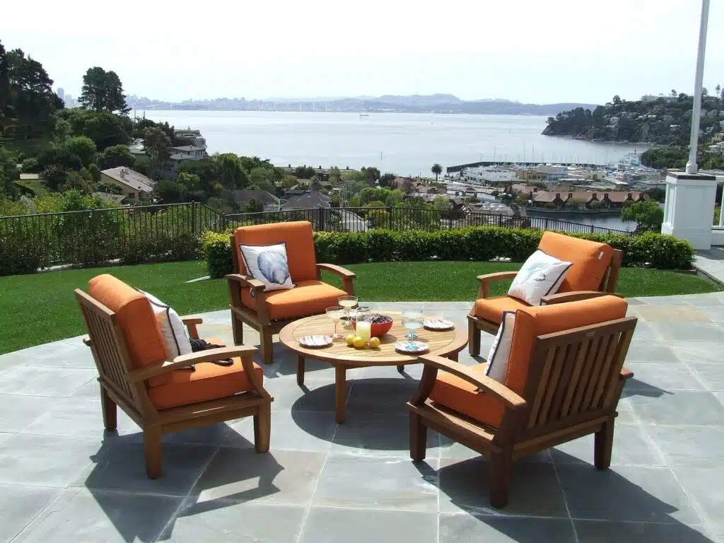 This image shows an outdoor patio or deck area with a scenic view of a body of water likely a bay or harbor surrounded by hills and trees The patio features several comfortable-looking wooden patio chairs with orange cushions arranged around a round wooden coffee table The table has a variety of dishes and fruits on it suggesting it is set up for an outdoor dining or relaxation area The patio is surrounded by lush greenery and has a tiled floor creating a peaceful and inviting atmosphere for enjoying the scenic waterfront view