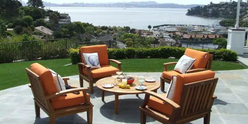 This image shows an outdoor patio or deck area with a scenic view of a body of water, likely a bay or harbor, surrounded by hills and trees. The patio features several comfortable-looking wooden patio chairs with orange cushions arranged around a round wooden coffee table. The table has a variety of dishes and fruits on it, suggesting it is set up for an outdoor dining or relaxation area. The patio is surrounded by lush greenery and has a tiled floor, creating a peaceful and inviting atmosphere for enjoying the scenic waterfront view.