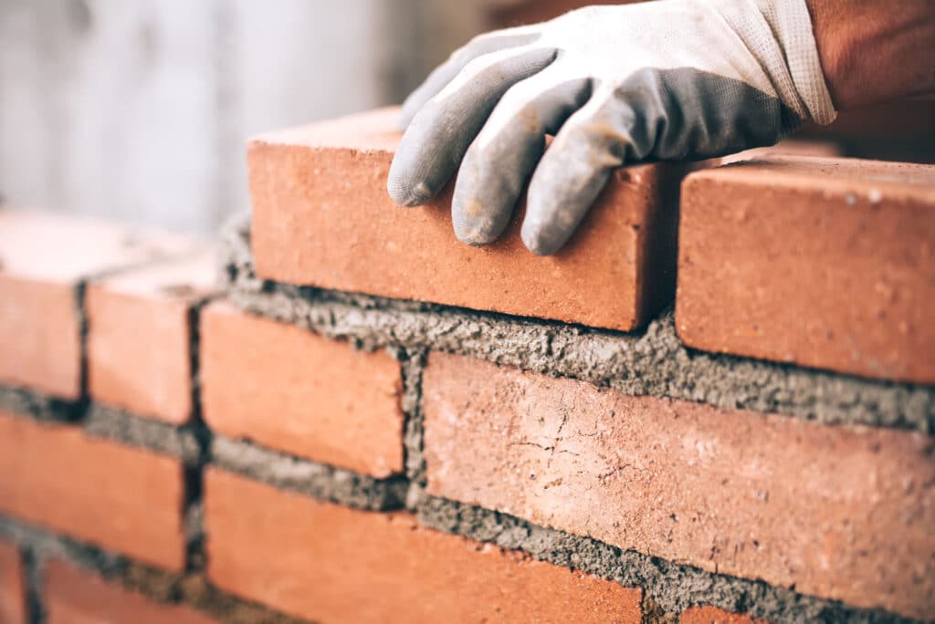 This image shows a close-up view of a persons hands wearing gloves while laying bricks The hands are carefully positioning a red clay brick on top of a layer of mortar creating a sturdy brick wall The bricks have a rough textured surface and the mortar between them is visible This image captures the skilled craftsmanship and attention to detail involved in the masonry work