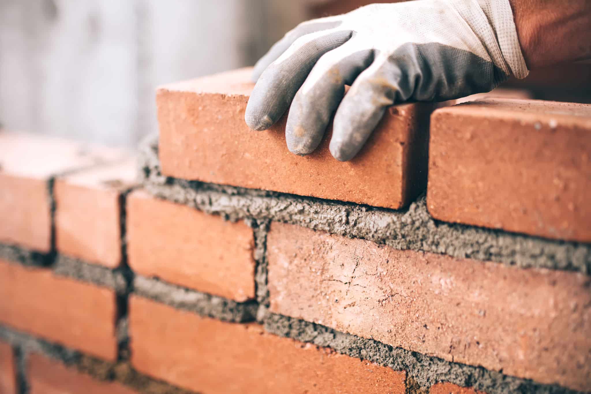 This image shows a close-up view of a person's hands wearing gloves while laying bricks. The hands are carefully positioning a red clay brick on top of a layer of mortar, creating a sturdy brick wall. The bricks have a rough, textured surface, and the mortar between them is visible. This image captures the skilled craftsmanship and attention to detail involved in the masonry work.