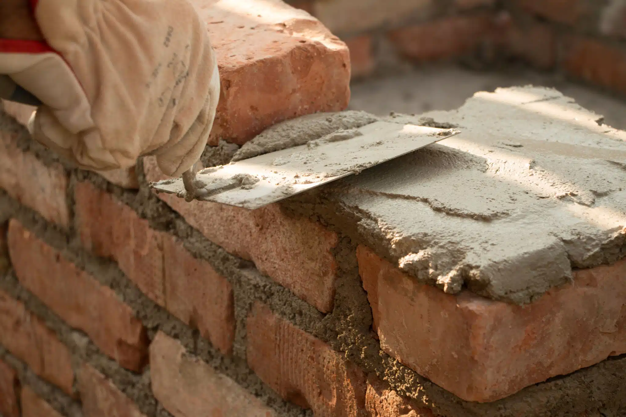 The image shows a close-up view of a masonry project It depicts several bricks and mortar with a trowel being used to apply the mortar between the bricks The bricks appear to be a reddish-brown color and the mortar is a greyish-white The texture and details of the bricks and mortar are clearly visible showcasing the craftsmanship involved in masonry work