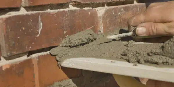 This image shows a close-up view of a persons hand applying mortar or cement between bricks during a masonry or bricklaying project The image captures the process of repairing or restoring a brick wall with the rough textured bricks and the wet gray mortar visible The focus is on the skilled hand work involved in the masonry repair highlighting the craftsmanship and attention to detail required for this type of construction work