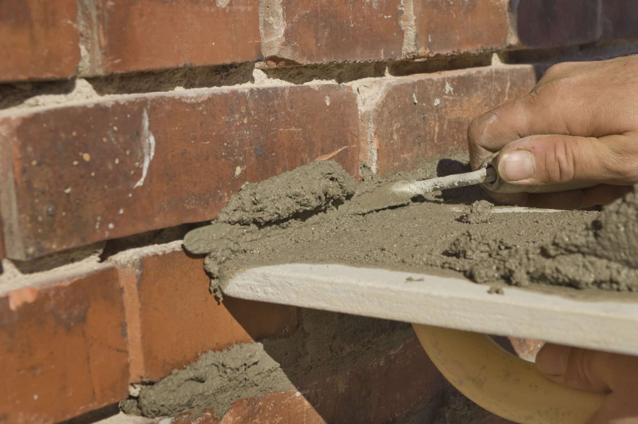 This image shows a close-up view of a persons hand applying mortar or cement between bricks during a masonry or bricklaying project The image captures the process of repairing or restoring a brick wall with the rough textured bricks and the wet gray mortar visible The focus is on the skilled hand work involved in the masonry repair highlighting the craftsmanship and attention to detail required for this type of construction work