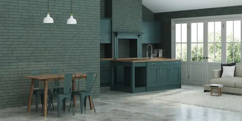 This image depicts a modern, open-concept kitchen and dining area. The space features a green brick wall, which contrasts with the light-colored concrete flooring. The kitchen cabinetry is painted in a deep, muted green color, providing a cohesive color scheme throughout the room. The kitchen island has a wooden countertop, and the room is illuminated by two white pendant lights. The dining area includes a wooden table and metal chairs, creating a rustic yet refined look. Large windows allow natural light to flood the space, creating a bright and airy atmosphere.