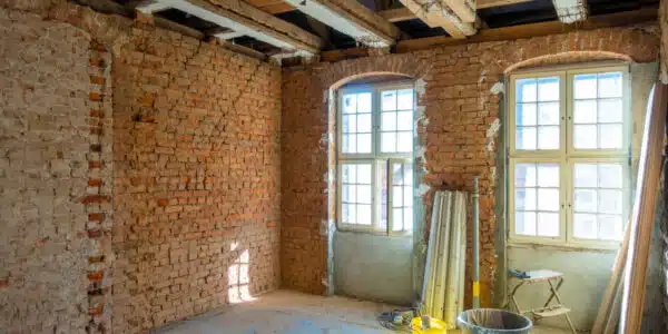 This image shows the interior of an old partially renovated building The walls are made of exposed brick with visible signs of wear and aging The room has large arched windows that let in natural light casting shadows on the floor Construction materials and tools such as a bucket and a stepladder are visible indicating that the space is currently undergoing renovation or restoration work The overall atmosphere conveys a sense of an unfinished yet charming historic space