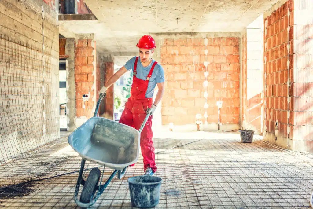This image shows a construction worker wearing a red hard hat and red overalls standing in a partially constructed building The worker is pushing a wheelbarrow filled with construction materials likely cement or mortar across the concrete floor of the unfinished interior The walls are made of exposed bricks and there is natural light coming in through openings in the structure The scene depicts an active construction site where the worker is engaged in the process of building or renovating the structure