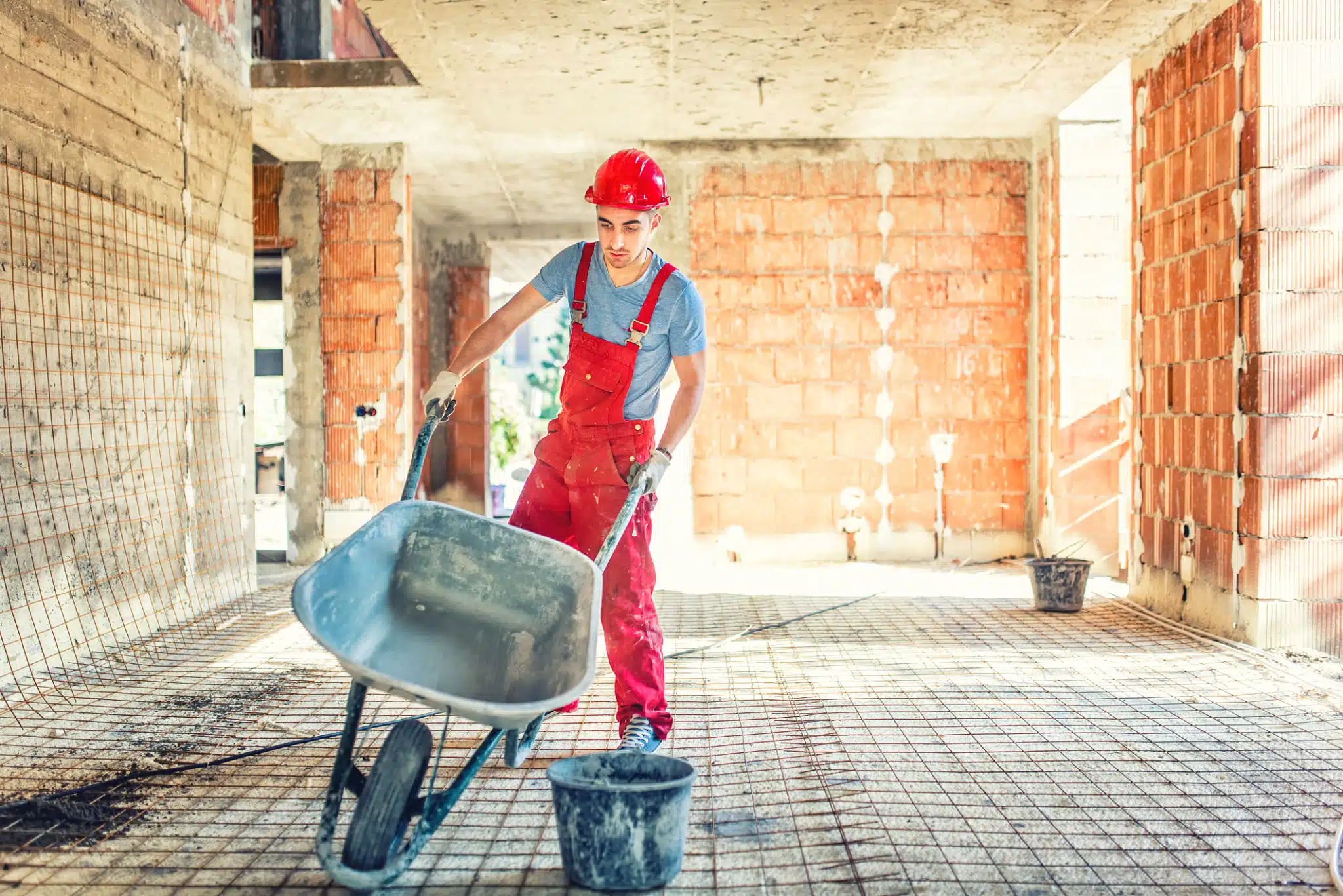 This image shows a construction worker wearing a red hard hat and red overalls, standing in a partially constructed building. The worker is pushing a wheelbarrow filled with construction materials, likely cement or mortar, across the concrete floor of the unfinished interior. The walls are made of exposed bricks, and there is natural light coming in through openings in the structure. The scene depicts an active construction site where the worker is engaged in the process of building or renovating the structure.