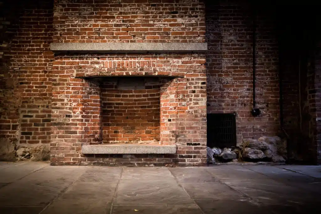 This image shows an old weathered brick wall with a recessed entryway or alcove The brickwork features a mix of red brown and dark shades creating a rustic textured appearance A stone or concrete ledge runs across the top of the recessed area providing a simple architectural detail The floor in front of the alcove appears to be paved with stone or concrete slabs A metal grate or vent can be seen in the lower right corner of the image adding to the industrial utilitarian feel of the scene