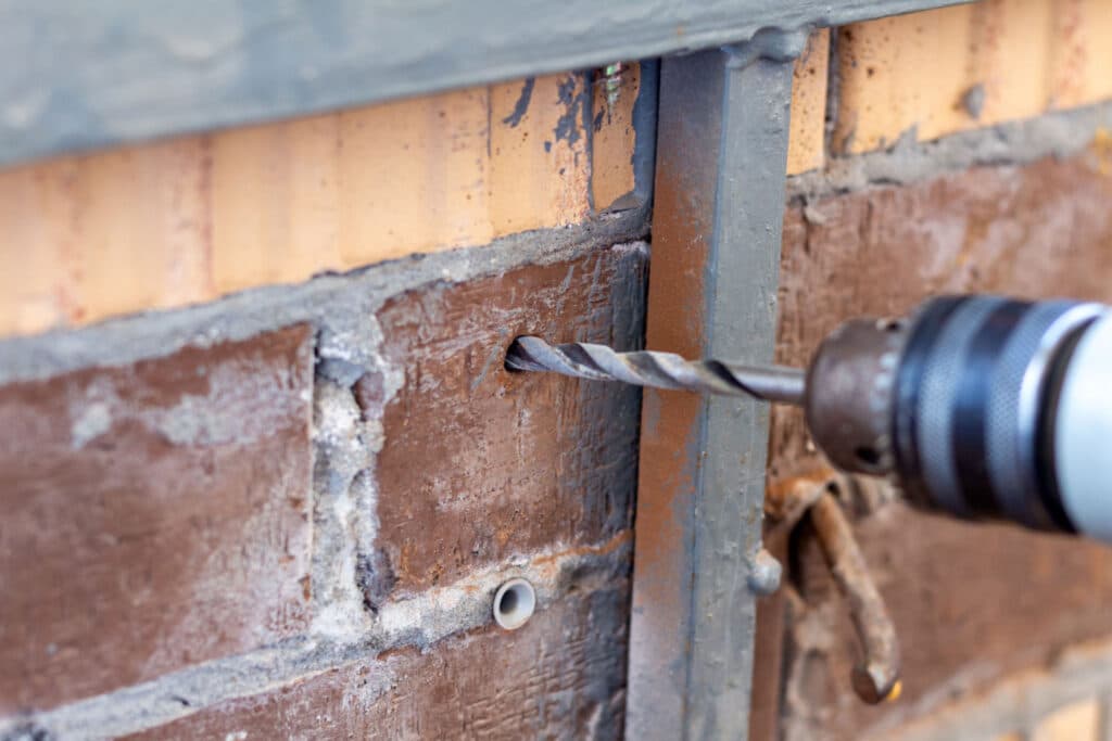 This image shows a close-up view of a brick wall undergoing repair or renovation The wall has areas of damaged and peeling plaster or stucco revealing the underlying brickwork A power drill with a drill bit is visible indicating that the person is likely drilling into the wall possibly to secure new materials or repair the damaged sections The image focuses on the textured weathered surface of the wall and the tools being used to address the issues