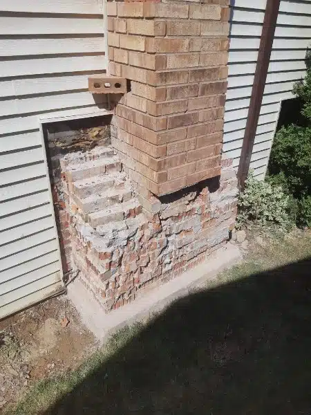 This image shows a brick chimney or column that appears to be partially damaged or deteriorating The bricks are a reddish-brown color and some of the mortar between the bricks has crumbled exposing the interior structure The chimney or column is situated next to a wooden fence or siding and there is some vegetation or grass visible in the foreground