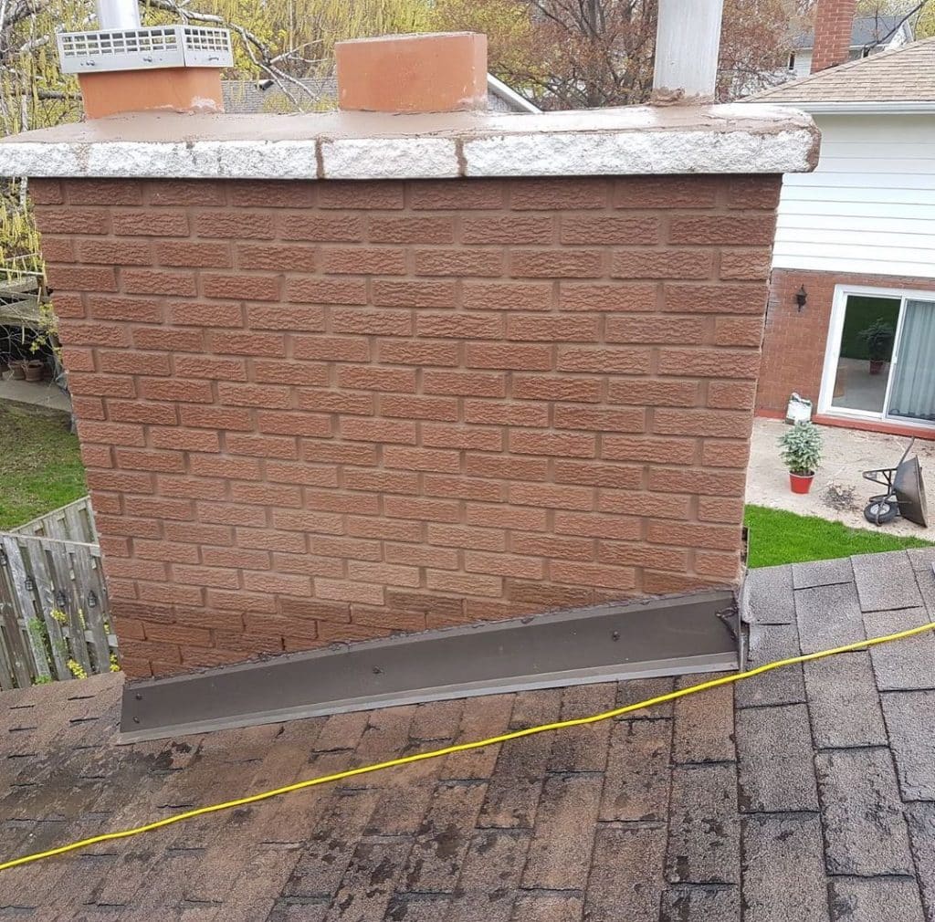 The image is a photo of a brick chimney on a shingled roof The chimney is made of red bricks and has a concrete cap on top There are two terra cotta chimney pots on top of the chimney The chimney is attached to a house with white siding and a red door There is a black metal flashing at the base of the chimney A yellow measuring tape is stretched across the roof in front of the chimney There is a backyard visible in the background with a wooden fence and patio furniture
