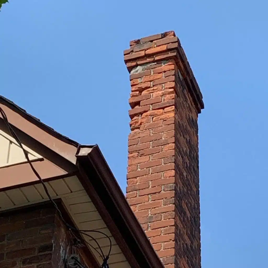 This image shows a close-up view of a brick chimney or chimney stack on the roof of a building The chimney is constructed with reddish-brown bricks and has a slightly irregular stacked appearance The sky in the background is a clear bright blue color providing a vivid contrast to the warm tones of the brickwork