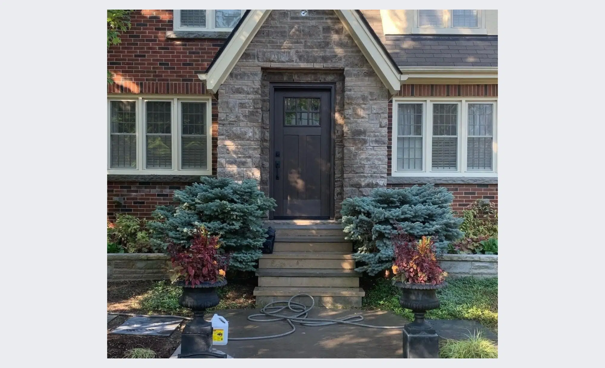 This image shows the front entrance of a residential home The home has a brick and stone exterior with a prominent black wooden front door framed by stone On either side of the front steps are large planters filled with lush blue-green evergreen shrubs The landscaping around the entrance includes additional plants and flowers creating a welcoming and well-maintained appearance The image also shows some tools and equipment on the front steps suggesting some ongoing work or maintenance being performed on the property