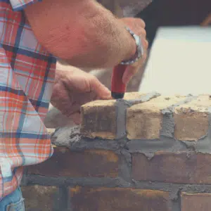 The image shows the hands of a worker wearing a plaid shirt as they use a trowel to apply mortar to a brick wall The worker is carefully spreading the mortar between the bricks demonstrating the skilled masonry work involved in constructing a brick structure