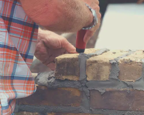 The image shows the hands of a worker, wearing a plaid shirt, as they use a trowel to apply mortar to a brick wall. The worker is carefully spreading the mortar between the bricks, demonstrating the skilled masonry work involved in constructing a brick structure.