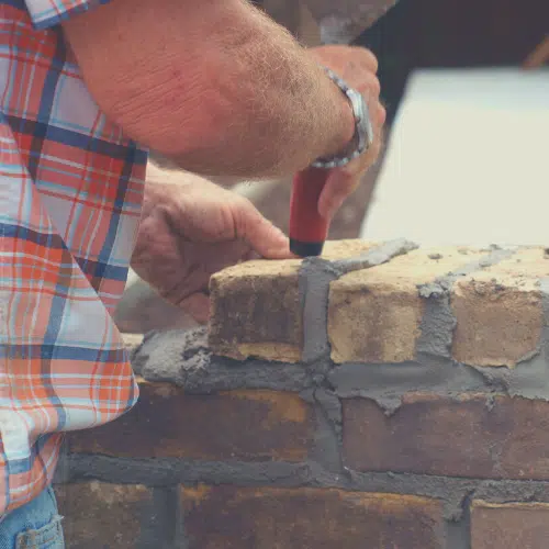 The image shows the hands of a worker, wearing a plaid shirt, as they use a trowel to apply mortar to a brick wall. The worker is carefully spreading the mortar between the bricks, demonstrating the skilled masonry work involved in constructing a brick structure.