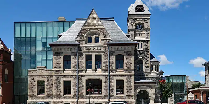 This image shows a historic stone building with a distinctive architectural style. The building has a large clock tower with a peaked roof, and the main structure features ornate stone detailing, arched windows, and a triangular pediment at the top. There are several glass-enclosed modern additions attached to the sides of the building, creating an interesting contrast between the old and new elements. The building is surrounded by a small landscaped area with flowers and benches, and there are a few cars parked in front of it, indicating this is likely a public or civic building located in an urban setting.