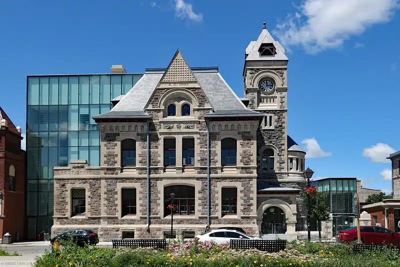 This image shows a historic stone building with a distinctive architectural style The building has a large clock tower with a peaked roof and the main structure features ornate stone detailing arched windows and a triangular pediment at the top There are several glass-enclosed modern additions attached to the sides of the building creating an interesting contrast between the old and new elements The building is surrounded by a small landscaped area with flowers and benches and there are a few cars parked in front of it indicating this is likely a public or civic building located in an urban setting