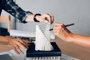 This image shows the hands of two people working on a model or architectural design made of wooden blocks The blocks are arranged in a stepped fan-like pattern and one person is using a pen or marker to make adjustments or markings on the model The other persons hands are visible suggesting they are collaborating on the design The image conveys a sense of creativity planning and attention to detail in the design process
