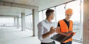 This image shows two individuals in a construction site or unfinished building One person is wearing a white shirt and appears to be an office worker or manager while the other person is wearing an orange safety vest and hard hat indicating they are a construction worker or technician The two individuals are engaged in a conversation likely discussing the progress or details of the construction project The image conveys a collaborative and professional interaction between the office worker and the construction worker within the unfinished industrial-looking environment