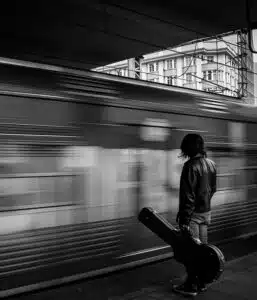 This image depicts a black and white scene of a person standing on a train platform as a train rushes by in the background The person is wearing a leather jacket and appears to be waiting or observing the passing train The motion of the train creates a blurred effect emphasizing the speed and movement The buildings in the background are visible but out of focus drawing the viewers attention to the solitary figure on the platform