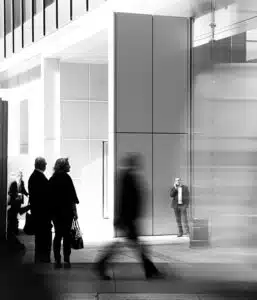This image appears to be a black and white photograph taken in a modern minimalist interior space The main focus is a large sleek refrigerator or appliance against a backdrop of glass walls and windows In the foreground there are several blurred figures likely people walking through the space The overall scene conveys a sense of movement and activity within a clean architectural environment