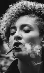 This black-and-white image depicts a close-up portrait of a woman with curly shoulder-length hair She has a piercing in her nose and is smoking a cigarette with smoke billowing from her lips The womans eyes are closed and she has a pensive introspective expression on her face The lighting and contrast create a dramatic moody atmosphere in the photograph