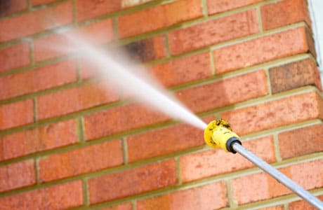 This image shows a yellow garden hose nozzle spraying water against a brick wall The brick wall has a reddish-orange color and a textured surface The water spray from the nozzle is visible creating a mist-like effect The image conveys the idea of cleaning or maintaining a brick structure using a water hose