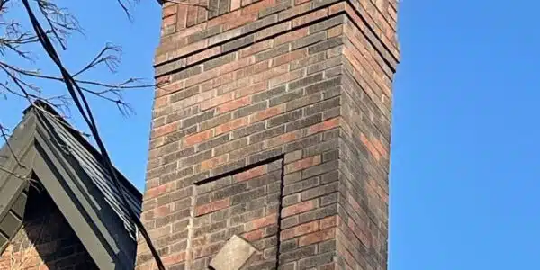 This image shows a tall brick chimney on a building The chimney is constructed with a mix of red and brown bricks arranged in a decorative pattern The chimney extends above the roofline and has a distinctive tapered shape at the top The sky in the background is a clear bright blue providing a striking contrast to the warm tones of the brick