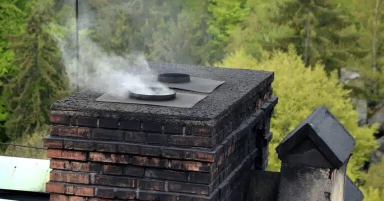 This image shows a brick chimney or outdoor fireplace with a smoky fire burning inside The chimney is constructed of weathered dark-colored bricks and has a flat square top with two metal cooking surfaces or burners Smoke is rising from the chimney indicating that the fire is active The chimney is situated in a forested green outdoor environment with trees and foliage visible in the background