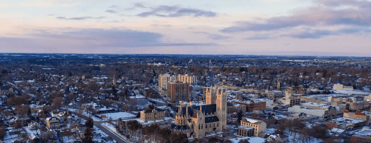 This image shows a panoramic view of a snowy cityscape at sunset The city appears to be a mid-sized urban area with a mix of modern high-rise buildings and historic architecture including a prominent cathedral or church with Gothic-style spires The city is surrounded by a winter landscape with trees and buildings covered in snow The sky is filled with colorful clouds creating a warm and picturesque atmosphere Overall the image conveys a scenic and atmospheric view of the city in the winter season