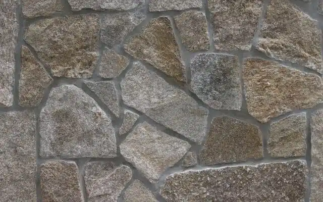 This image shows a close-up view of a stone wall or patio surface. The wall is constructed using irregularly shaped natural stones in various shades of gray, brown, and beige. The stones are arranged in a random, mosaic-like pattern, creating an interesting and textured surface. The stones appear to be a mix of different types of natural stone, such as granite or slate, giving the wall a rustic and earthy appearance.