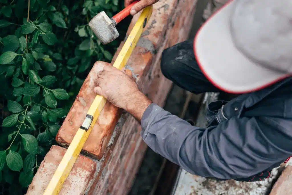 A person using a yellow spirit level to check the level of the bricks on a red brick wall. The person is wearing a grey jacket, blue jeans and a white hard hat. The mortar is white and there is a green plant in the background
