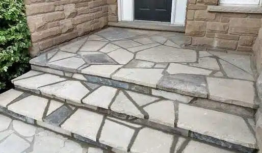 This image shows a stone patio or entryway in front of a building The patio is made of irregularly shaped stone tiles or slabs in various shades of gray creating a unique and natural-looking pattern The stones are arranged in an irregular mosaic-like design with some larger stones and smaller pieces filling in the gaps There is a set of stone steps leading up to the patio and the overall look is rustic and earthy blending well with the stone exterior of the building