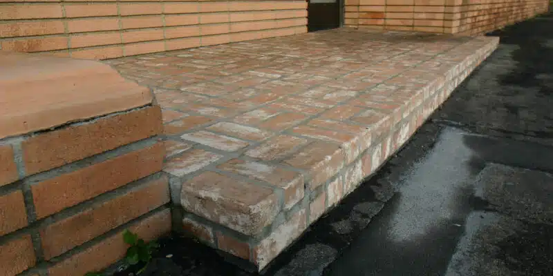 This image shows a brick walkway or patio leading up to a brick building. The walkway is made of rectangular brick pavers arranged in a pattern, with some gaps between the bricks. The bricks have a reddish-brown color and appear to be weathered, with some discoloration and texture visible. The walkway leads up to a set of brick steps that provide access to the building's entrance. Overall, the image depicts a well-constructed masonry feature that is part of the building's exterior design.