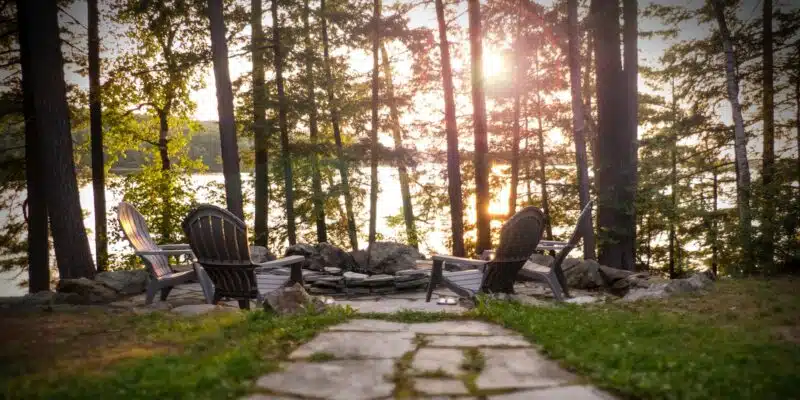 This image depicts a serene outdoor setting in a wooded area near a lake or body of water. The scene features several Adirondack-style wooden chairs arranged on a stone patio or path, surrounded by lush greenery and tall pine trees. The warm, golden light of the setting sun filters through the trees, creating a peaceful and tranquil atmosphere. The overall image conveys a sense of relaxation and connection with nature.